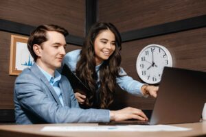 Two professionals reviewing time zone coordination on laptop with clock displayed, representing nearshore team collaboration and time management across distributed locations.