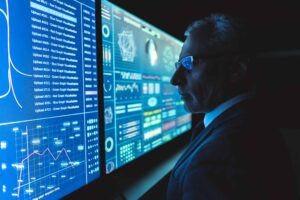 Software development manager monitoring risk dashboards and real-time analytics across multiple displays to identify and track project threats and KRIs.